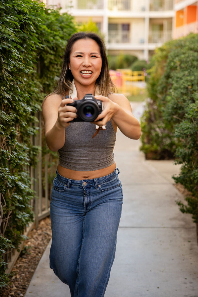 A smiling photographer walks toward the camera holding a DSLR, framed by green hedges along an outdoor walkway, representing a Bay Area lifestyle photographer at work.
