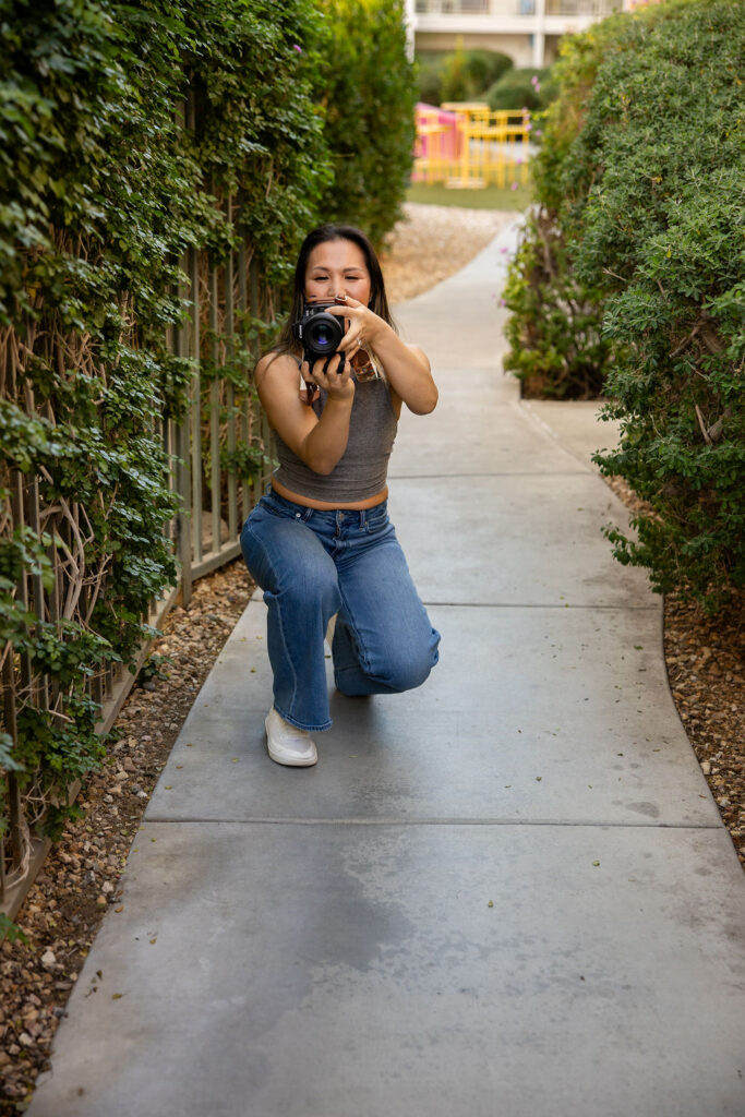 A woman kneels on a garden walkway while holding a camera up to her face, capturing photos outdoors in natural light as a lifestyle photographer serving Bay Area families