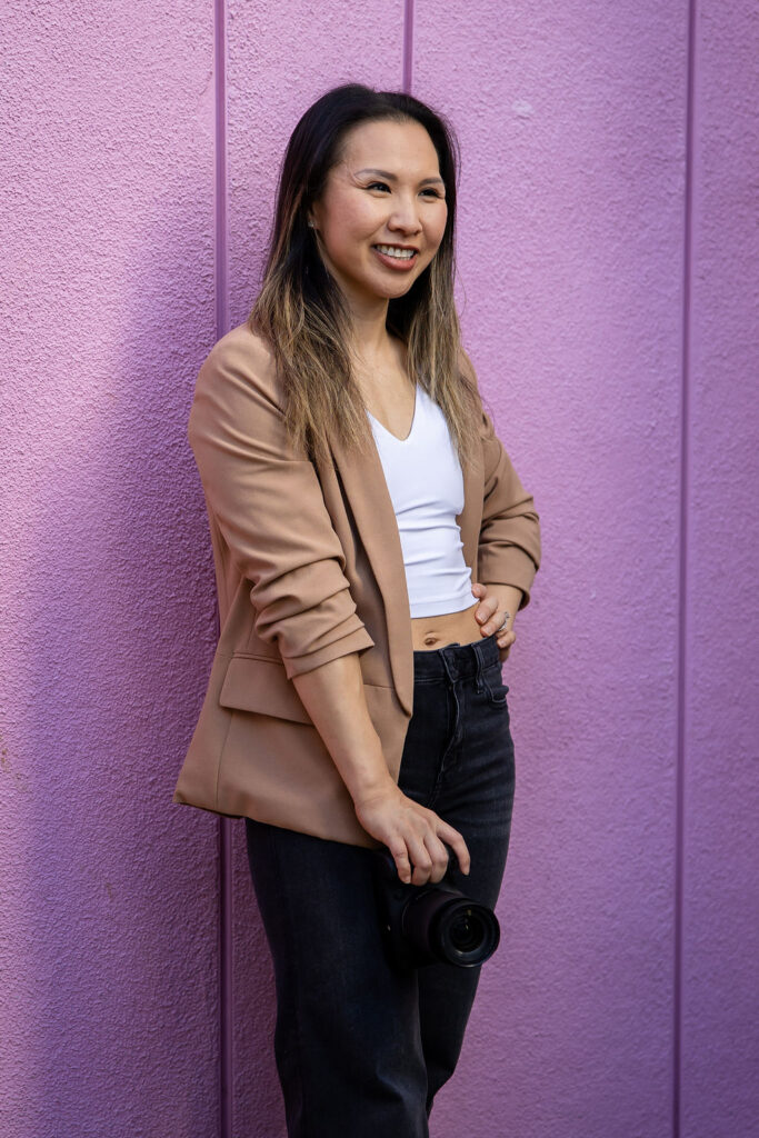 A fun photographer stands against a purple wall holding a camera at her side, smiling softly in natural light in the San Francisco Bay Area.