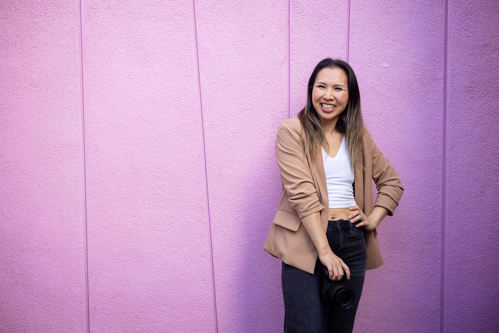 A photographer stands slightly off center against a wide pink wall, holding her camera and smiling in an open relaxed stance.