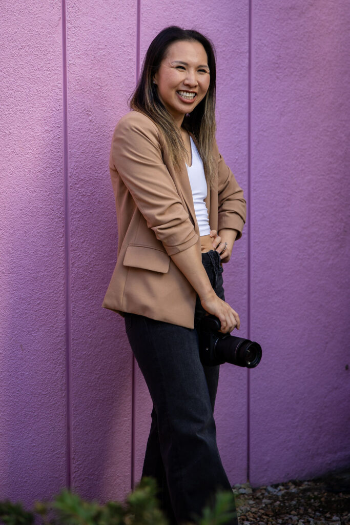 A natural light photographer stands angled toward the camera with her hands relaxed, smiling gently against a purple wall in the Peninsula.