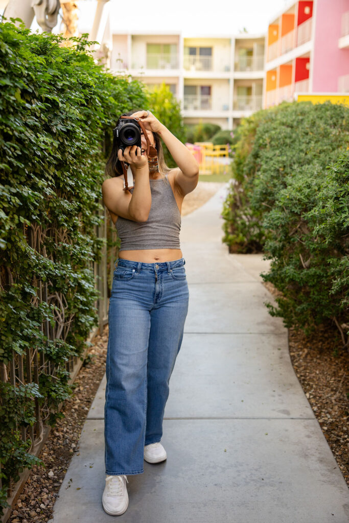 A lifestyle photographer walks down a garden path while lifting her camera to take a photo, capturing real life moments as a Bay Area photographer.