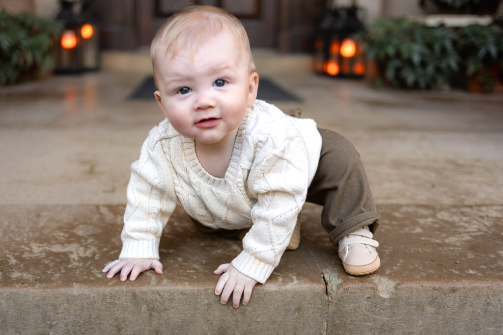A baby crawls forward on a stone step outdoors, looking directly at the camera with soft light and warm tones around him.