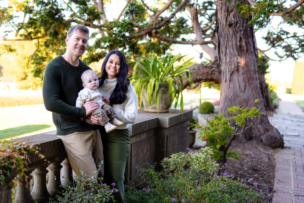 A family stands near a stone railing surrounded by plants, holding their baby while looking upward during a Lifestyle Photography session outdoors.