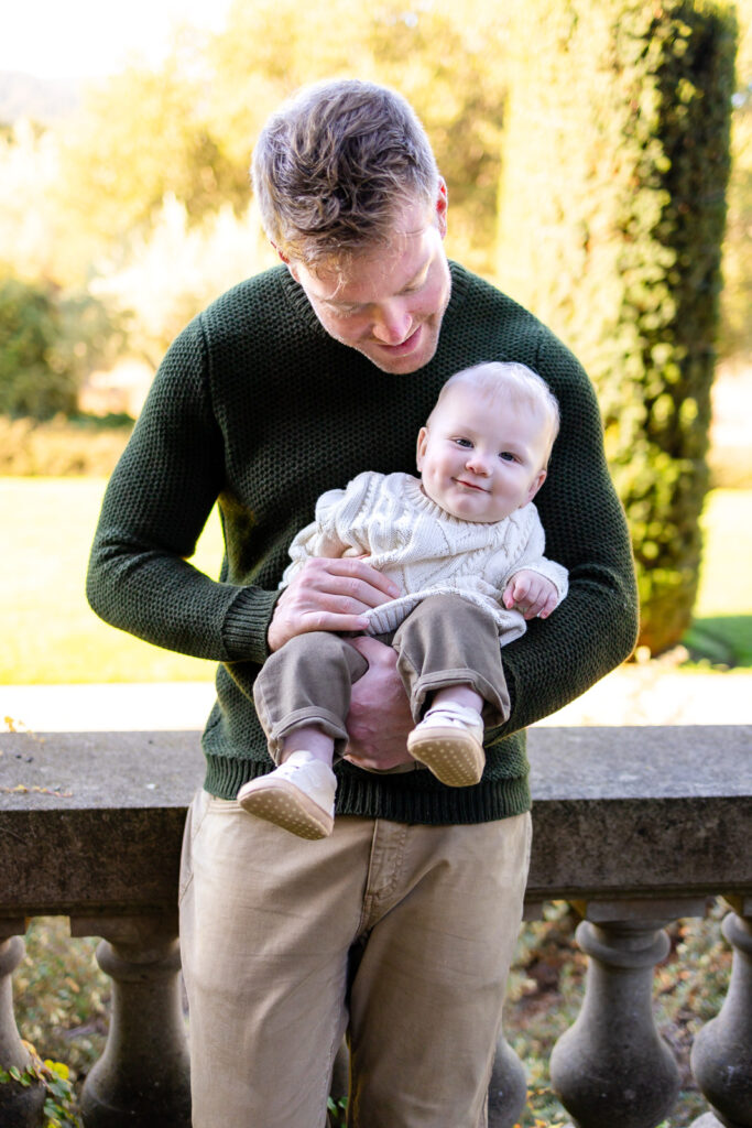 A father holds his baby at chest height while looking down with a gentle smile as the baby looks toward the camera.