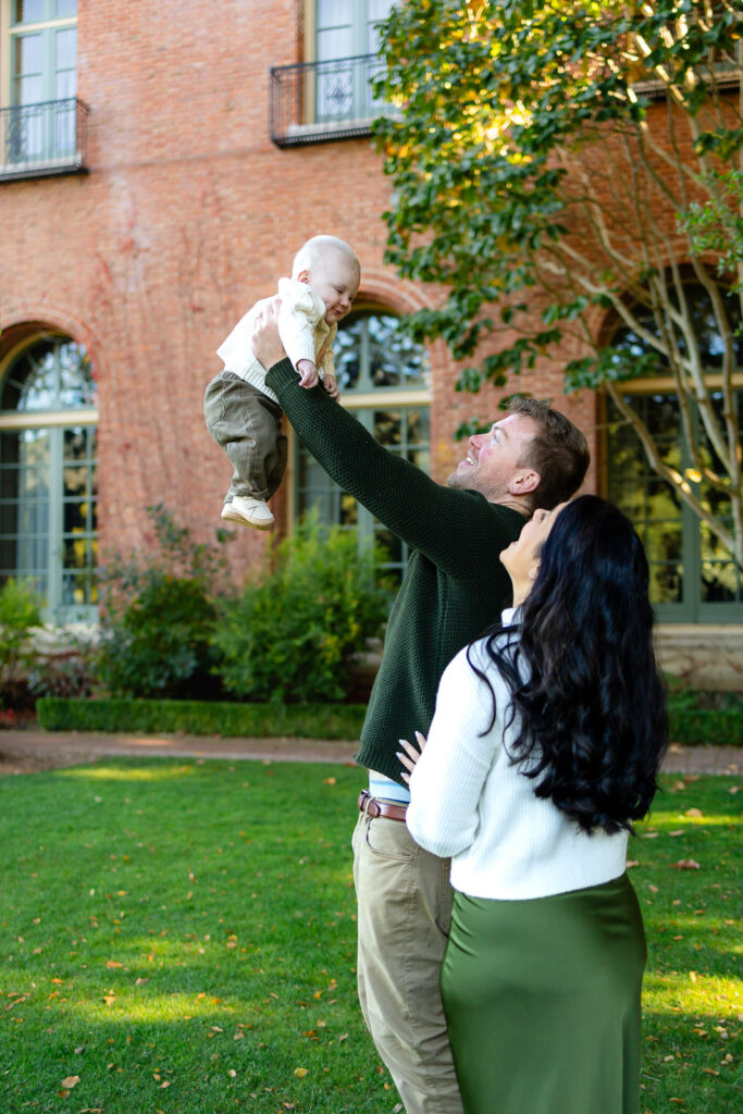 A father lifts his baby into the air while the mother looks on, smiling in a grassy courtyard during a Lifestyle Photography session.