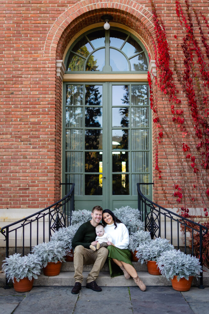 A family sits together on stone steps in front of a brick building, holding their baby and leaning close to one another.