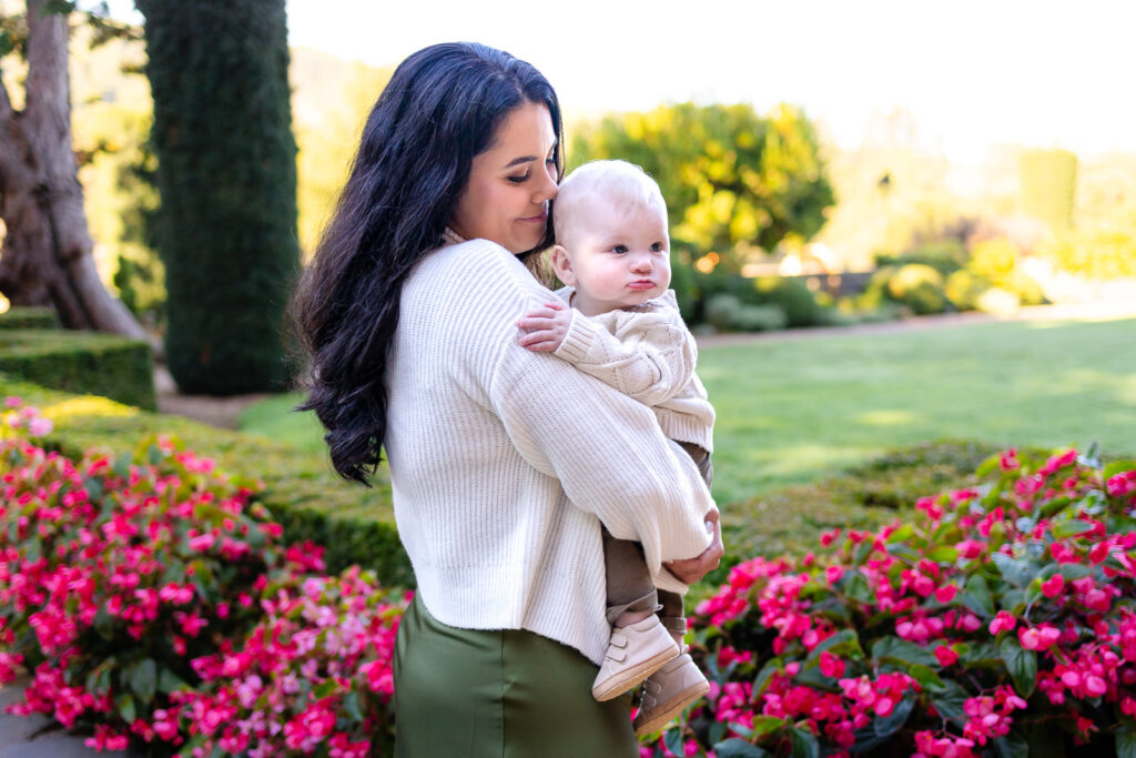 A mother holds her baby over her shoulder while standing near bright pink flowers in a landscaped garden.