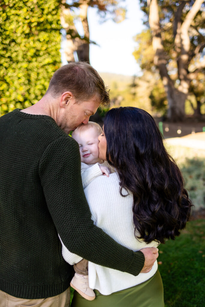 Parents lean in to kiss their baby on the cheeks while holding them close during a Lifestyle Photography session outdoors.