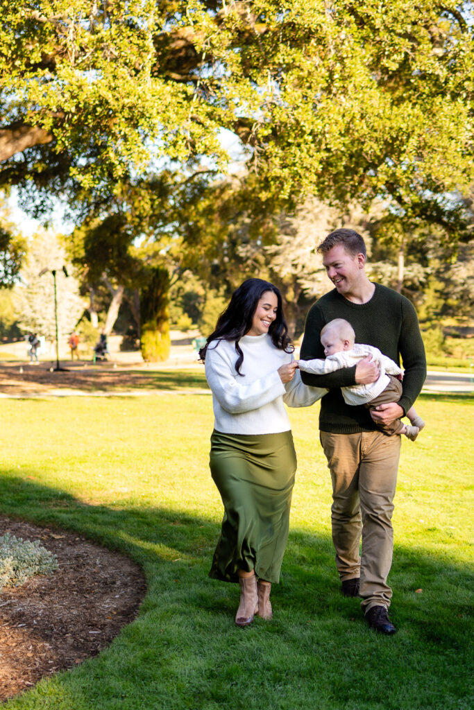 A family walks across a grassy lawn while the mother gently holds the baby’s hand as the father carries the child.