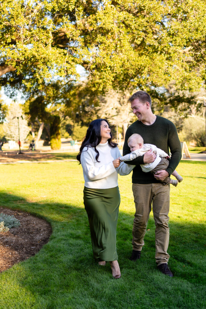 A couple walks side by side across green grass while sharing a smile as the father holds their baby.