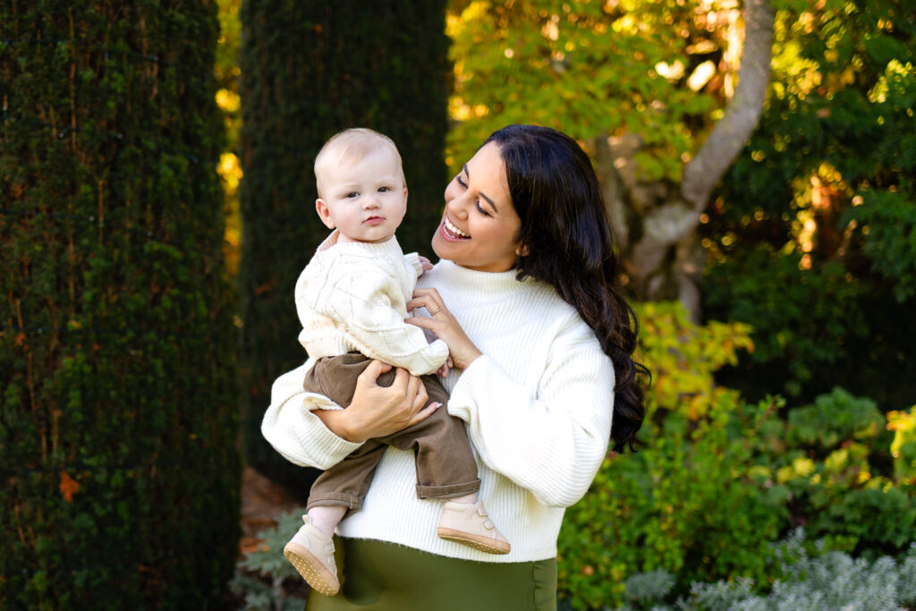 A mother holds her baby in her arms while smiling down at them, surrounded by lush greenery in soft afternoon light.