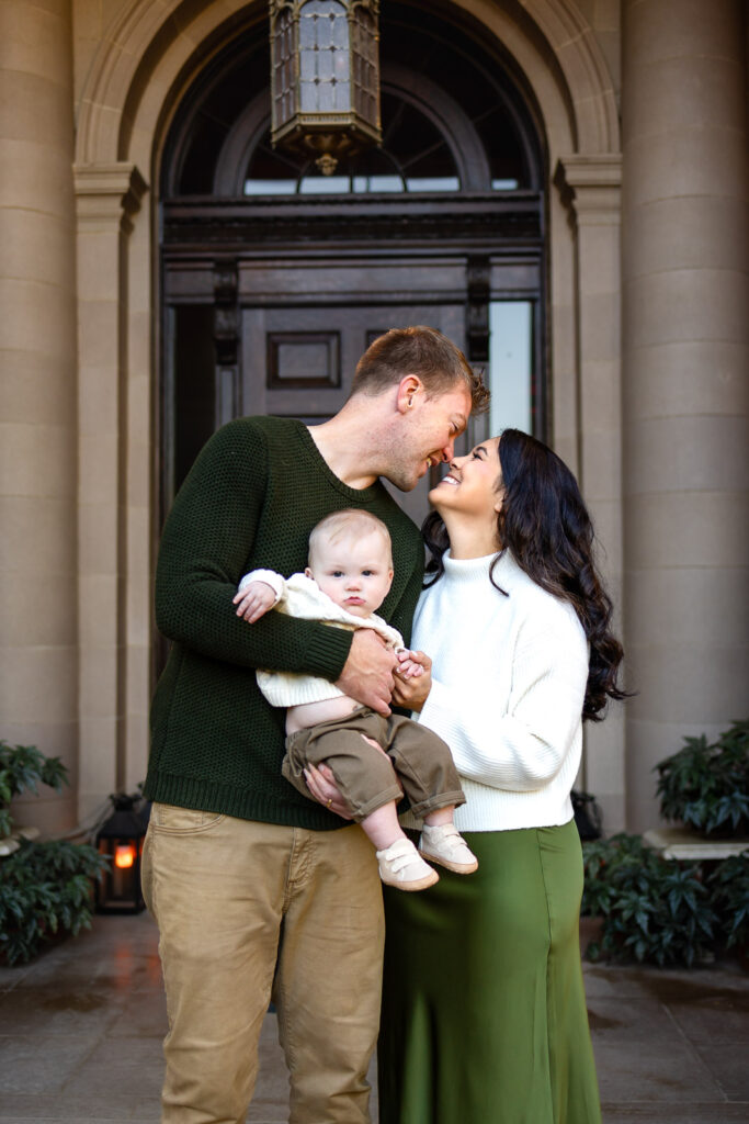 Parents stand close together holding their baby in front of a grand doorway, leaning in toward each other during a Lifestyle Photography session.