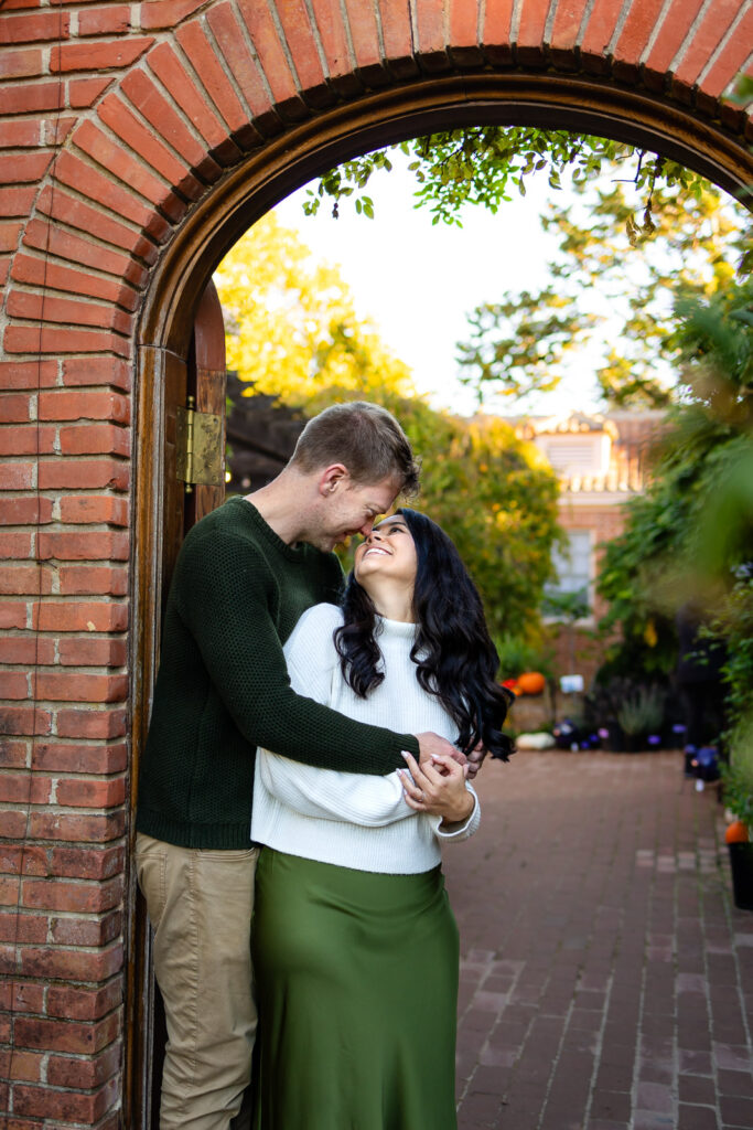 A couple embraces under a brick arch as the woman smiles upward, captured during a relaxed Lifestyle Photography session outdoors.