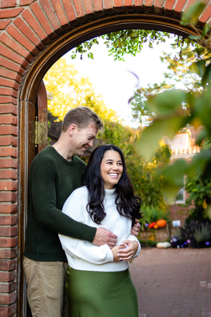 A couple laughs together as they stand under an archway, framed by soft greenery and autumn light from behind.