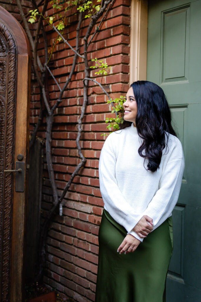 A woman stands by a brick wall and green door, looking off to the side with a gentle smile in natural light.