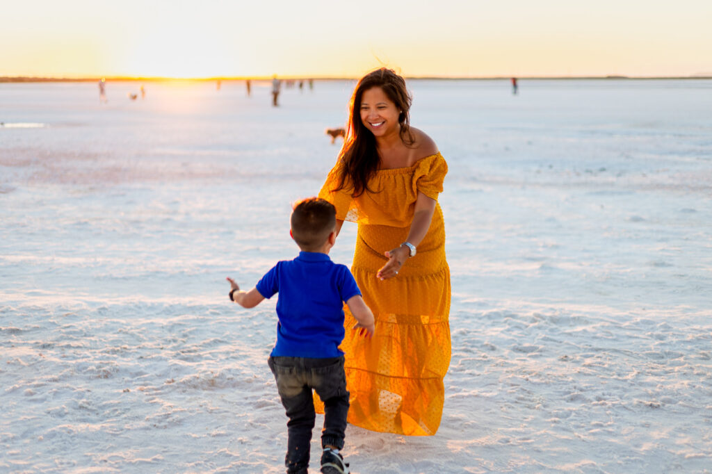 A mother in a flowing yellow dress opens her arms as her young son runs toward her across the beach at sunset.