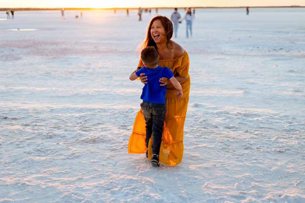 A mother laughs joyfully as her young son runs into her arms on a wide sandy beach during golden hour.