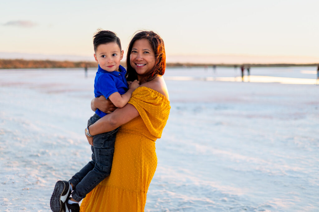 A mother holds her son close on the beach while both look toward the camera with soft smiles as the sun sets behind them.