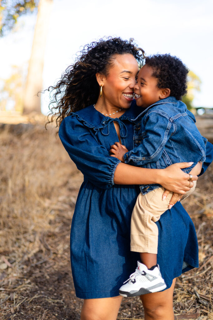 A mother holds her smiling toddler close as they laugh together outdoors during a Mommy and Me Photoshoot.
