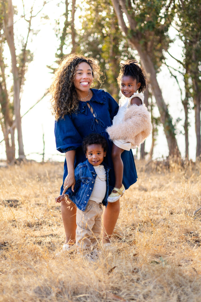 A mother stands in a grassy field holding one child while another stands close beside her, all smiling softly.