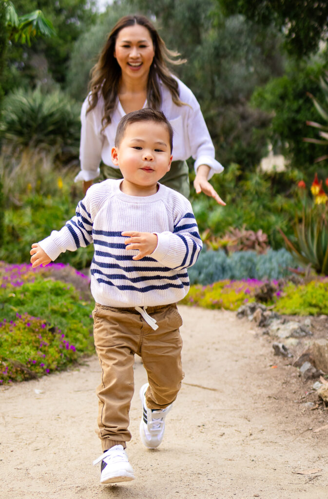 A toddler runs toward the camera on a garden trail while his mother follows behind smiling and reaching out.