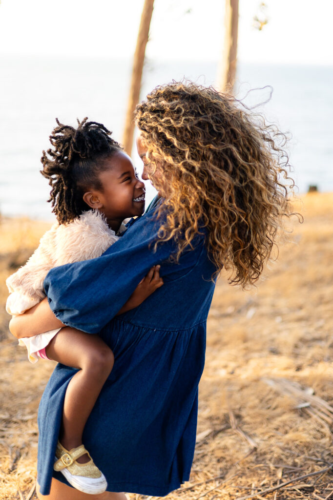 A mother lifts her daughter and they smile nose to nose while standing in tall grass near the water for a mommy and me session