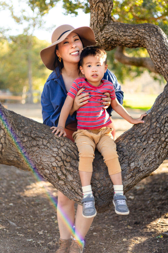 A mother smiles as she helps her young son sit on a large tree branch while standing close behind him in a sunlit park.
