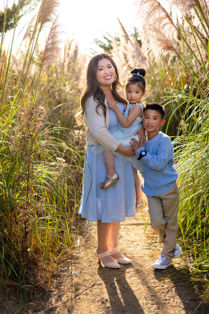 A mother stands holding her daughter while her son leans in beside them, all three posing together on a natural trail during a Mommy and Me Photoshoot.