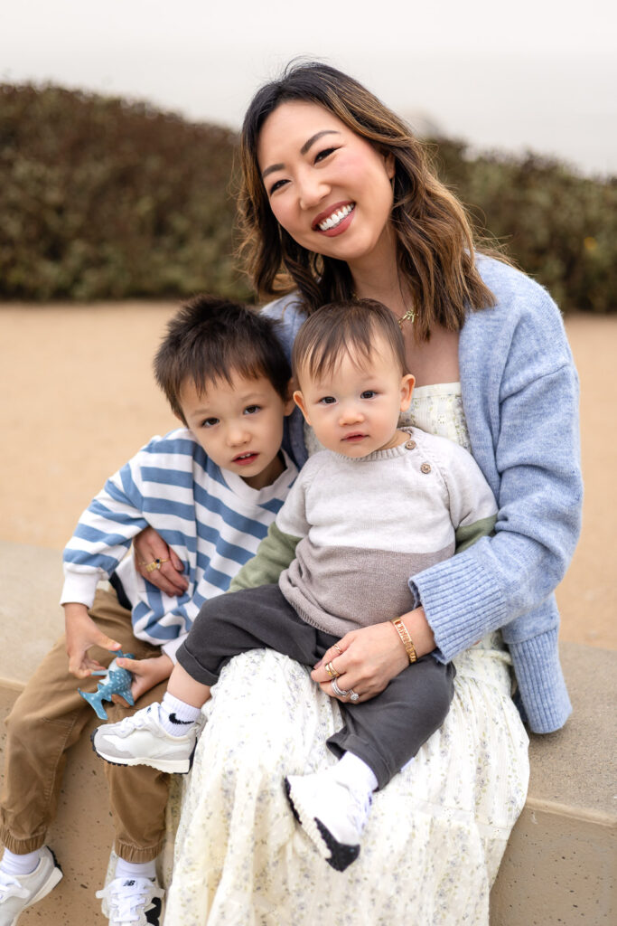 A mother sits on a low wall with her two young children gathered closely on her lap, all facing the camera outdoors during mommy and me mini session