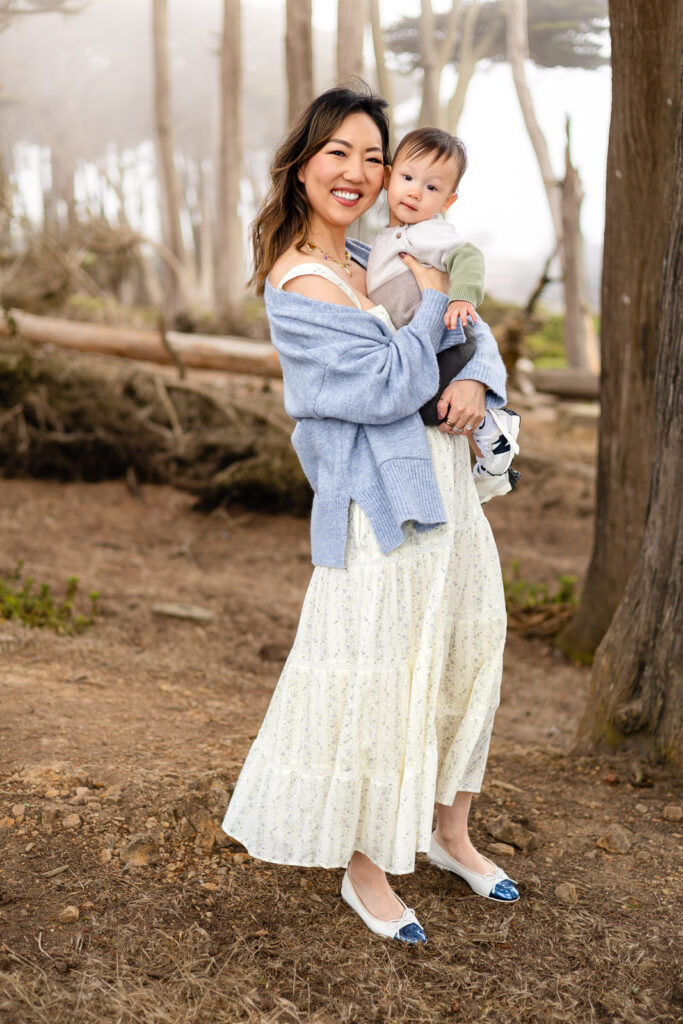 A mother holds her baby on her hip while standing among trees, smiling warmly toward the camera during mommy photoshoot