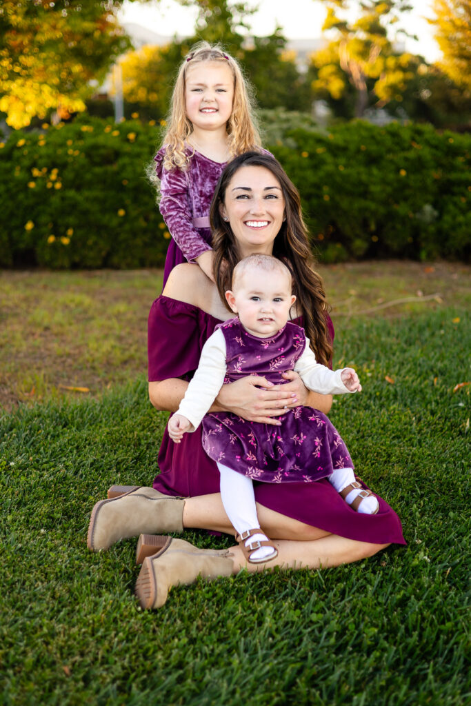 A mother sits on the grass holding her baby while her older daughter stands behind her with her hands resting on moms shoulders.