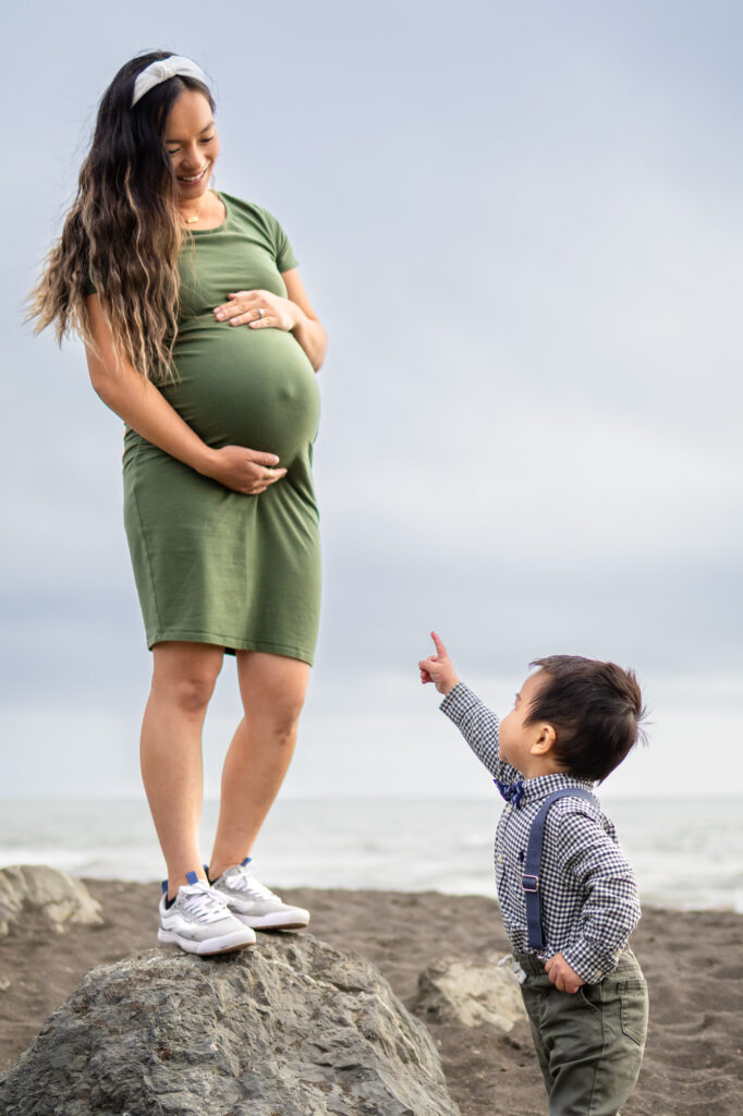 A pregnant mother stands on rocks at the beach as her toddler points up at her belly, creating a tender Mommy and Me Photoshoot moment.