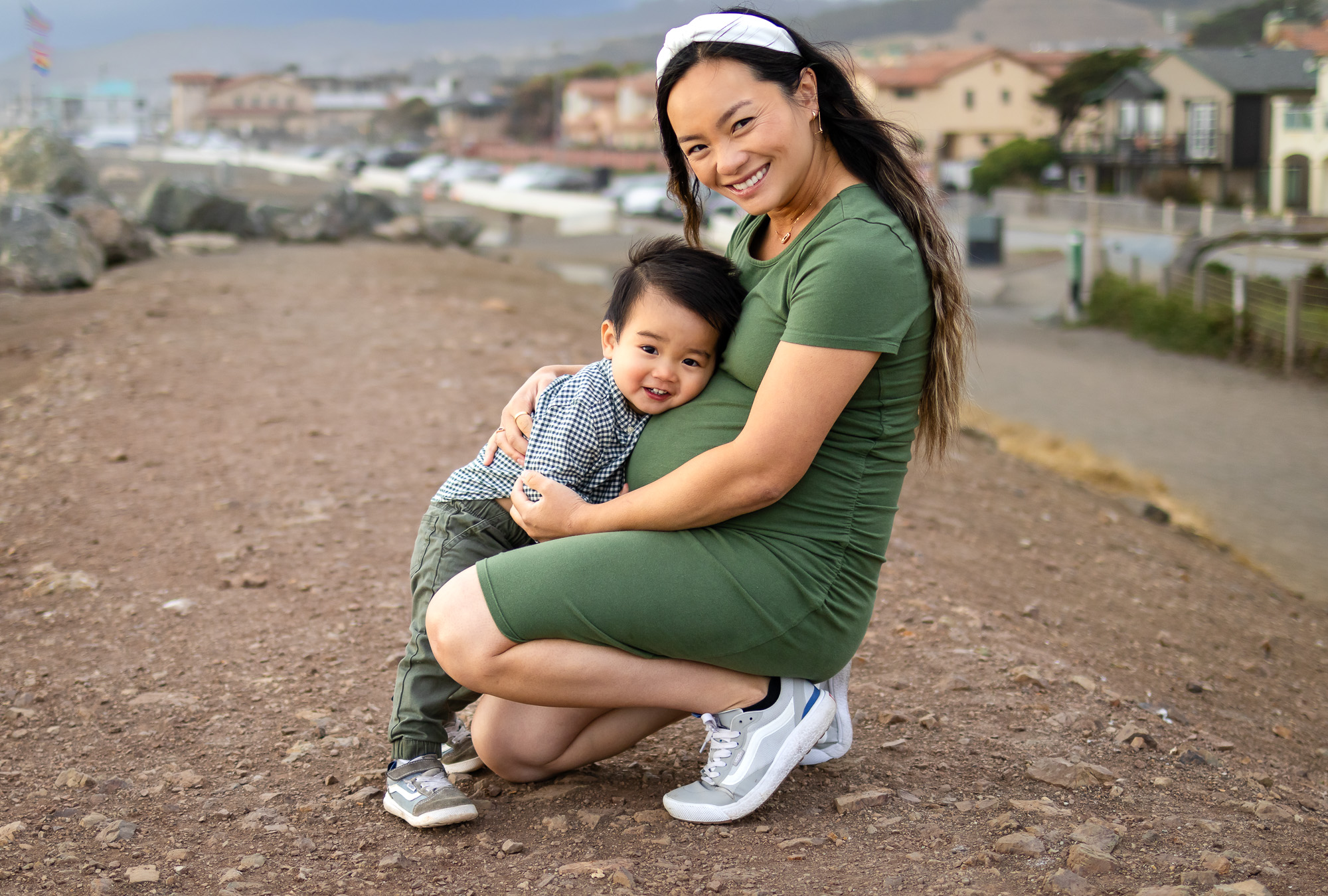 A mother crouches and hugs her smiling toddler close beside her belly on a seaside path during a Mommy and Me Photoshoot.