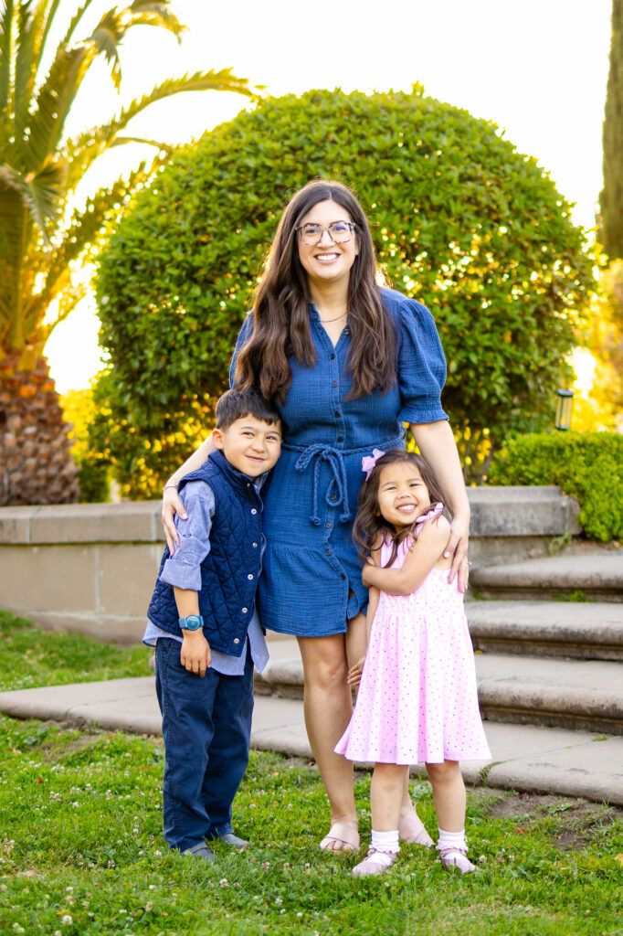 A mother stands with her two young children in a garden, all three smiling and leaning into each other.
