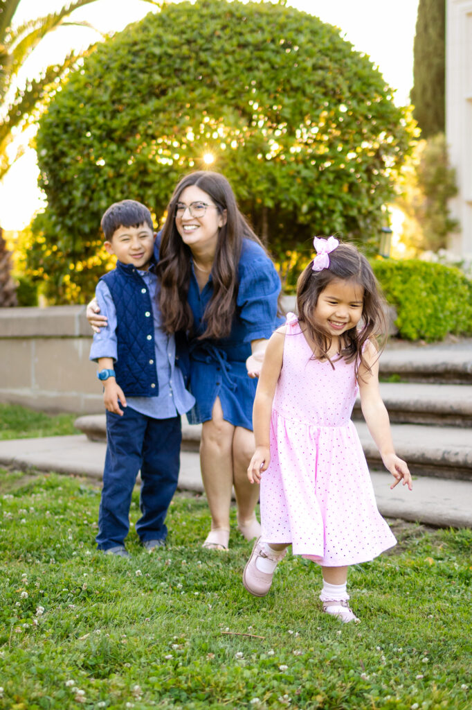 A mother gently guides her children forward on the grass as they walk and laugh together in soft evening light.