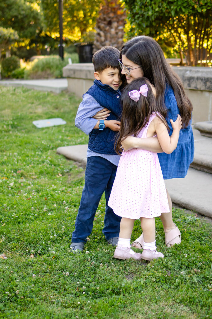 A mother kneels to hug her two children tightly as they wrap their arms around her in a group embrace.