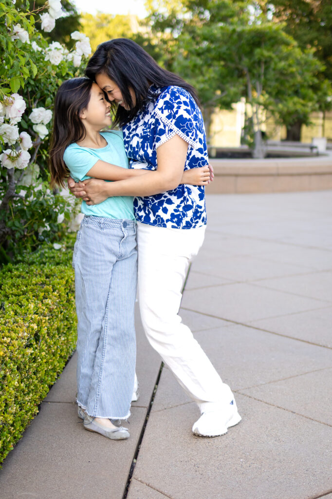 Mommy and me images of a mother and daughter stand close with their foreheads touching, smiling softly beside blooming white flowers.
