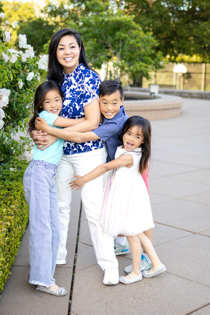 A mother stands surrounded by her three children as they hug her waist and smile toward the camera during a Mommy and Me Photoshoot.