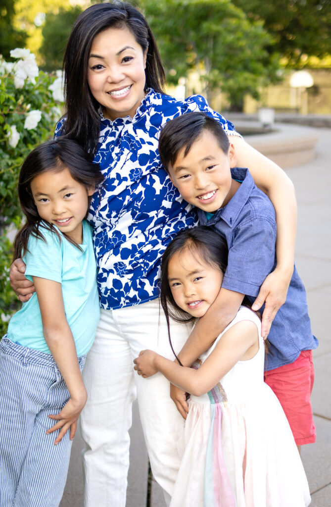 A mother crouches slightly as her children lean into her arms, all smiling together during a relaxed Mommy and Me Photoshoot outdoors.