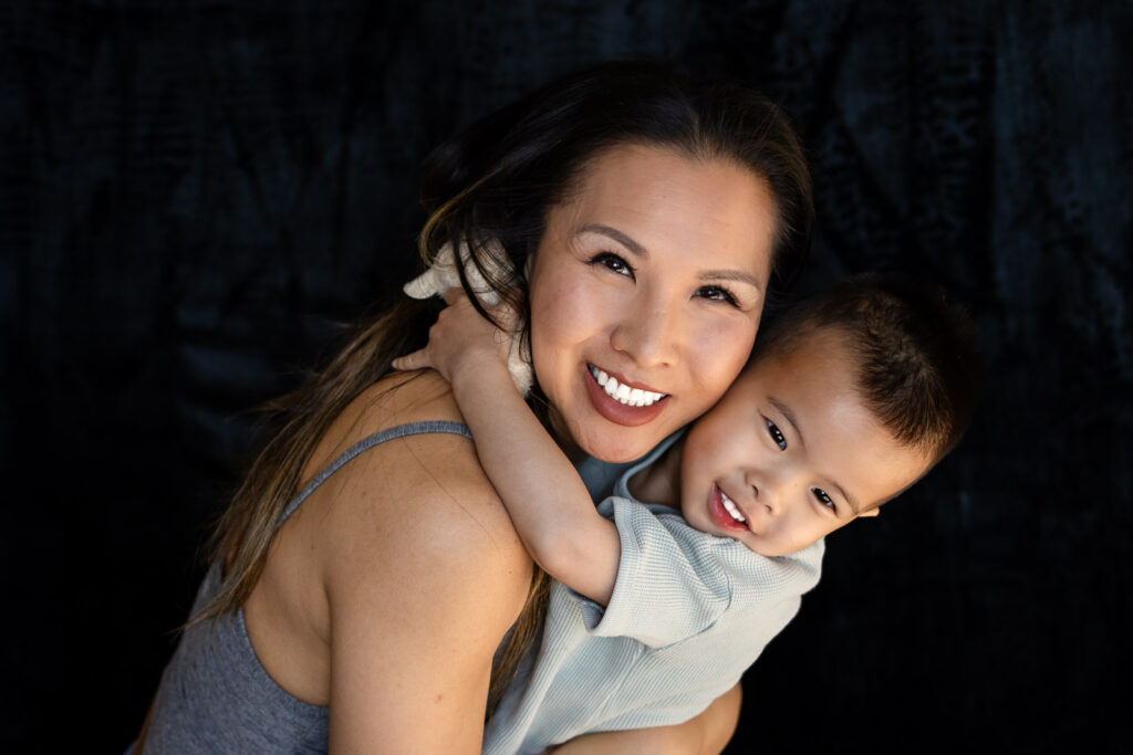 A mother holds her toddler close as he wraps his arms around her neck, both smiling softly against a dark studio backdrop during Mommy and child photoshoot