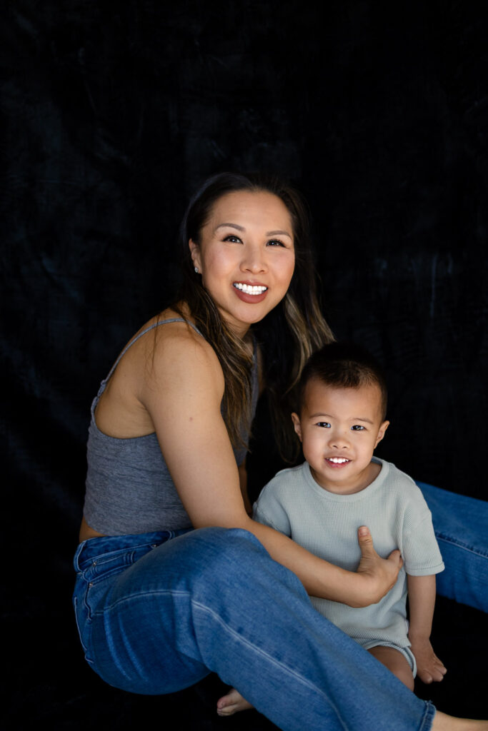 A mother sits on the floor beside her toddler, smiling warmly as he looks ahead with a happy relaxed expression during Motherhood Sessions