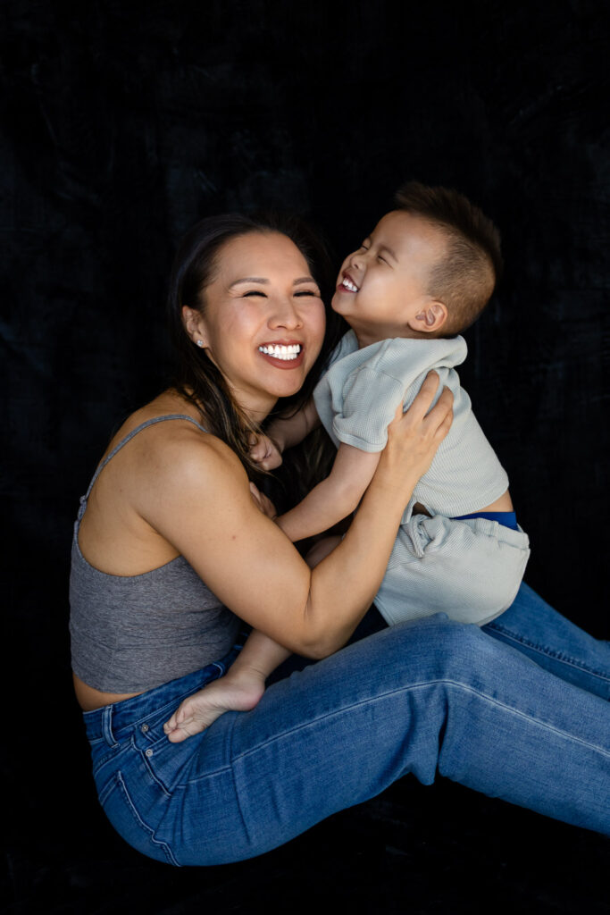 A mother sits on the floor smiling as her toddler leans in close, both laughing during a playful cuddle during Motherhood Sessions
