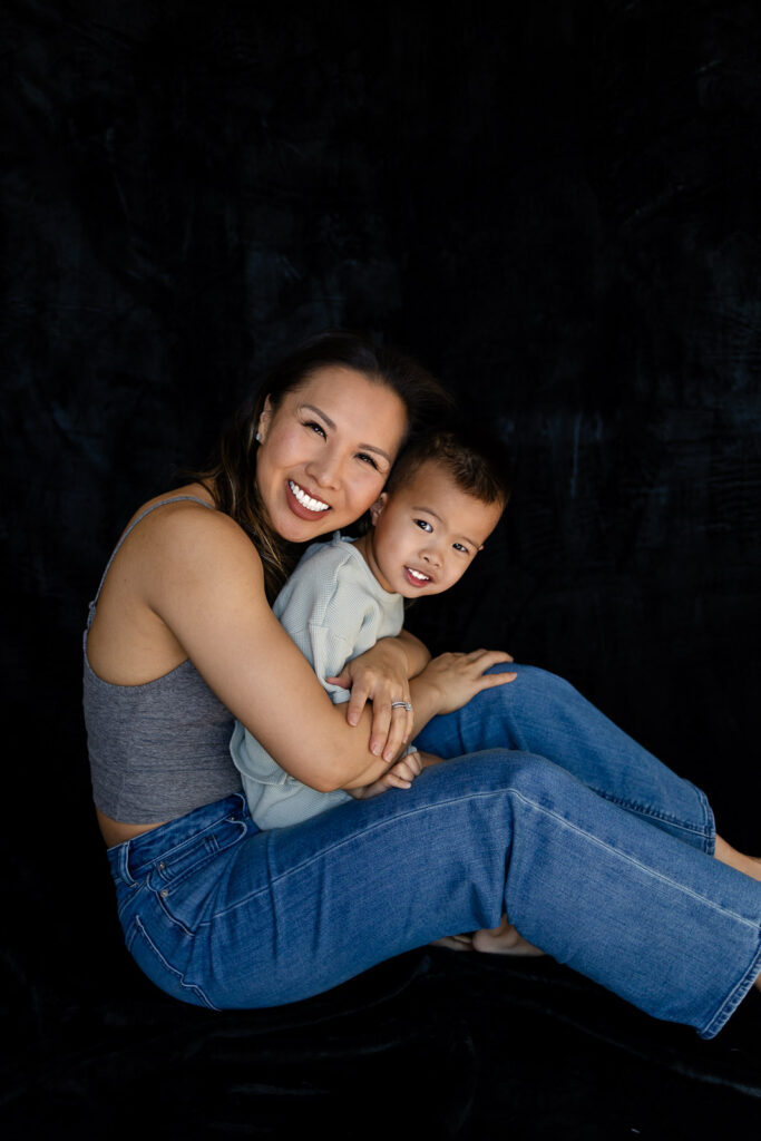 A mother wraps her arms around her toddler from behind as they sit side by side and smile softly at the camera during Mom and Kids Mini Session