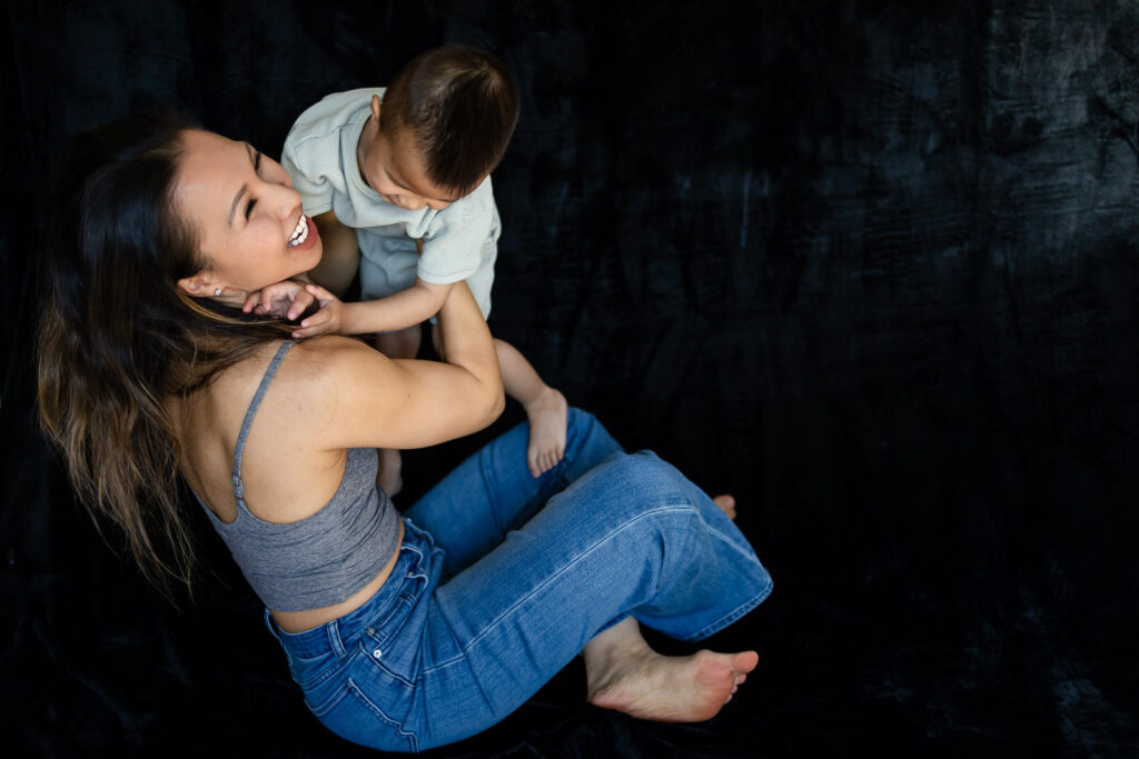 A mother looks up laughing as her toddler climbs toward her shoulders during a playful studio moment during Mom and Kids Mini Session