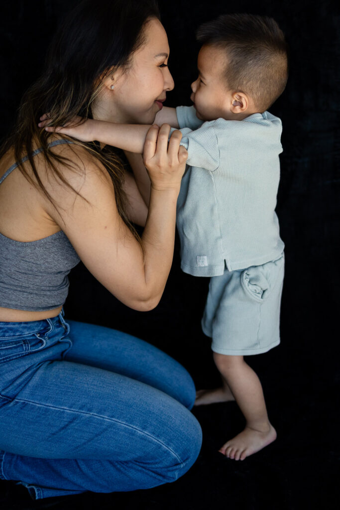 A mother kneels in front of her standing toddler as they hold each other’s arms and smile face to face during Mini Motherhood Portraits.