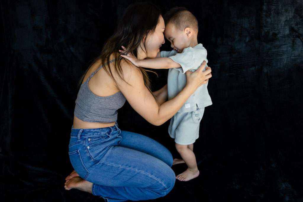 A mother kneels in front of her standing toddler as they hold each other’s arms and smile face to face.