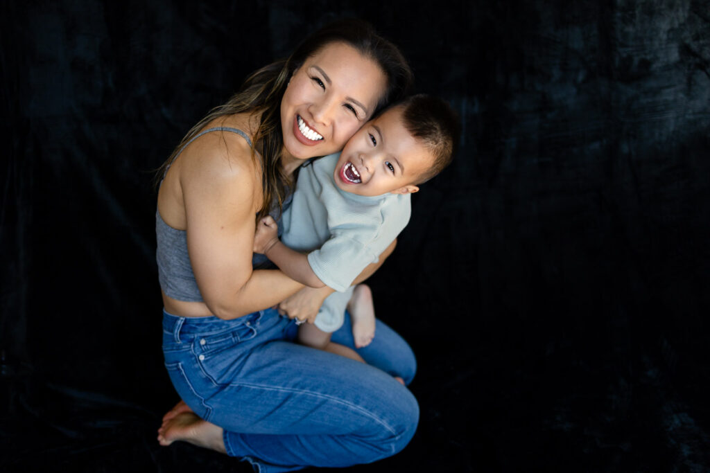 A mother kneels and hugs her toddler tightly as he laughs with his mouth open in a joyful embrace during Motherhood Mini Sessions.