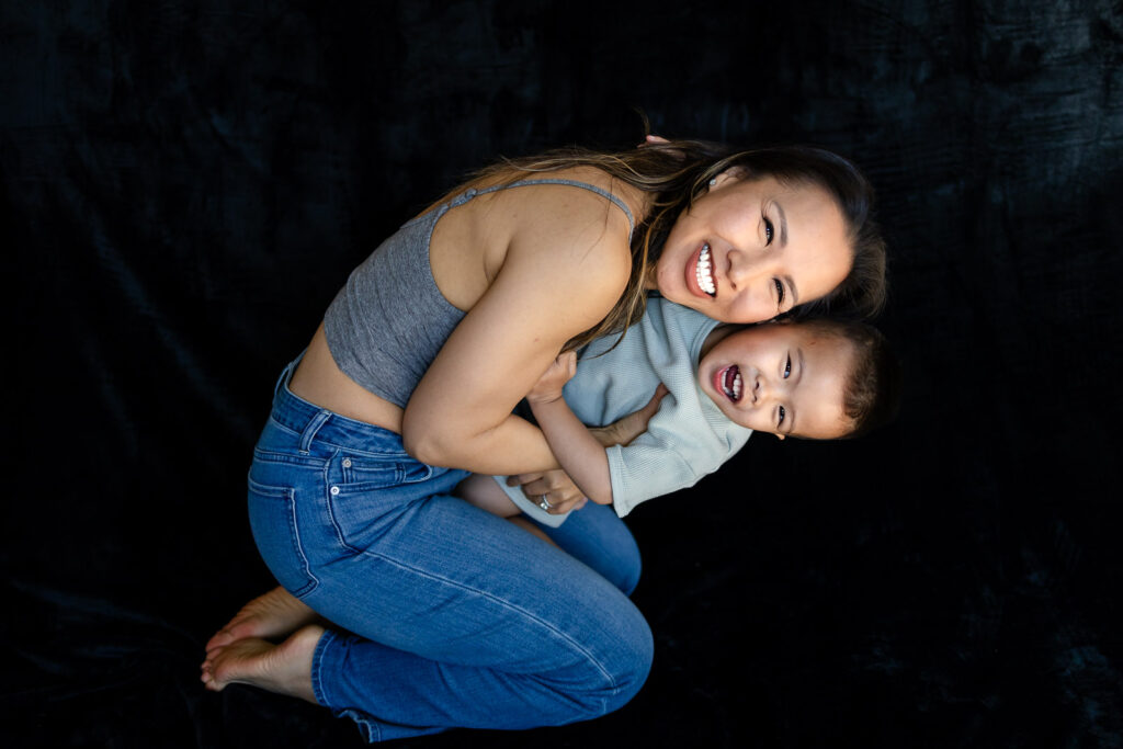 A mother crouches and hugs her toddler close as both laugh and tilt toward the camera during Mini Motherhood Portraits