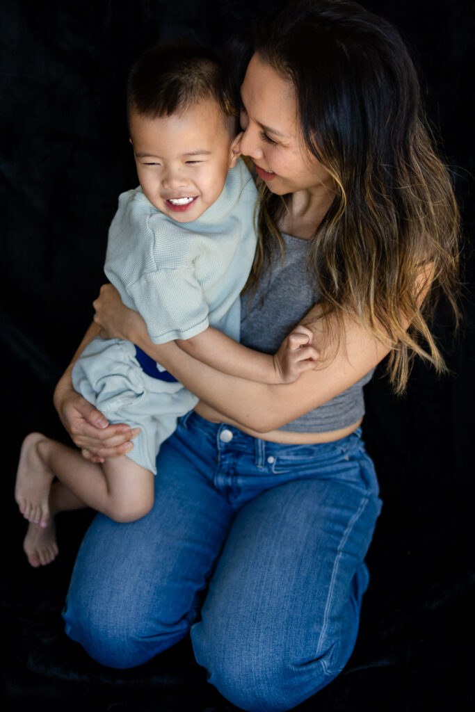 A mother sits holding her toddler on her lap while looking down at him with a calm affectionate smile.